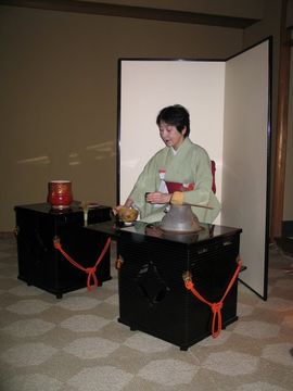 A woman holding a natsume performs the Ryū-rei ceremony.  Visible from far left to right are the red fresh water container (its lid is on the tana), the tea whisk, the tea bowl, the iron pot and the ladle (resting on the pot).