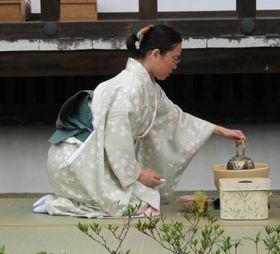 A woman wearing a kimono performs a tea ceremony outdoors, while seated in seiza position.